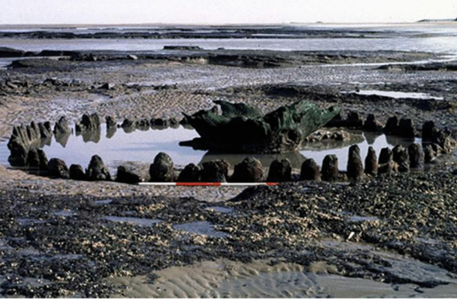 Seahenge, an ancient timber circle on the Norfolk coast, partially submerged in tidal waters. The structure consists of a central upturned tree stump surrounded by a ring of wooden posts