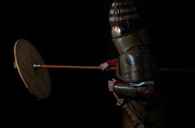 Participant wearing armor and hitting a shield while holding a spear.