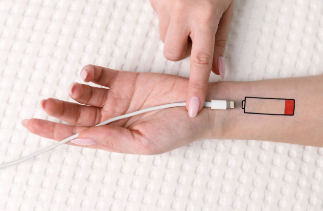 Low battery symbol drawn on human hand. Tired woman lying on the bed, trying to charge from USB, holding cable with your finger. Overwork, exhausted, chronic fatigue, living energy concept.