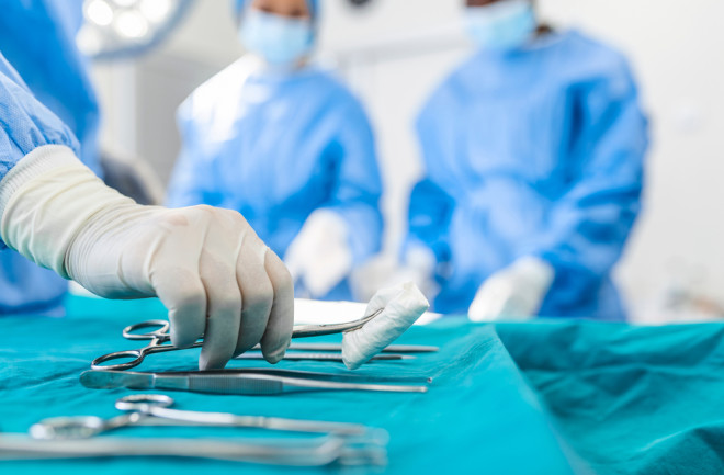 Nurse hand taking surgical instrument for group of surgeons at background operating patient in surgical theatre. Steel medical instruments ready to be used. Surgery and emergency concept