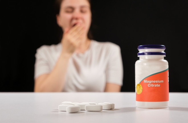 A tired woman sits at a table with a bottle of magnesium citrate and several tablets in front of her